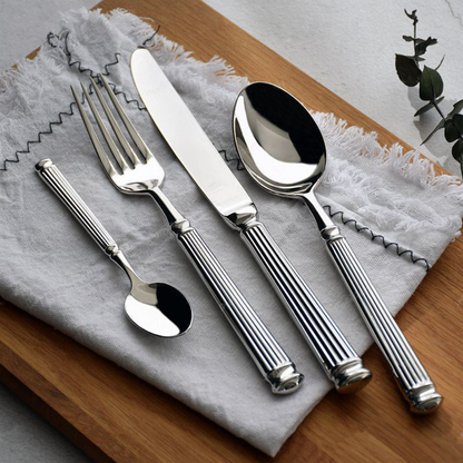 Five pieces of fluted stainless steel cutlery fanned across grey linen on a wooden board, the handles ending in stepped rounded caps