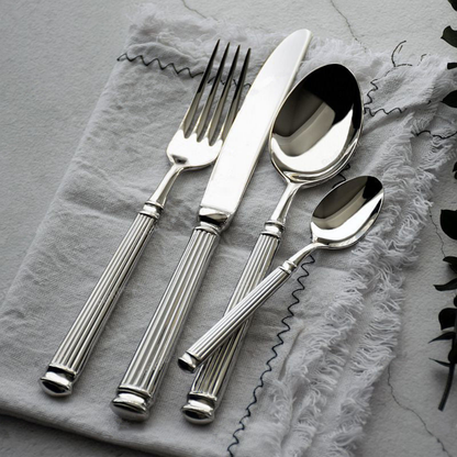 Five pieces of mirror polished cutlery on crumpled grey linen over marble, the fork tines and spoon bowls in view