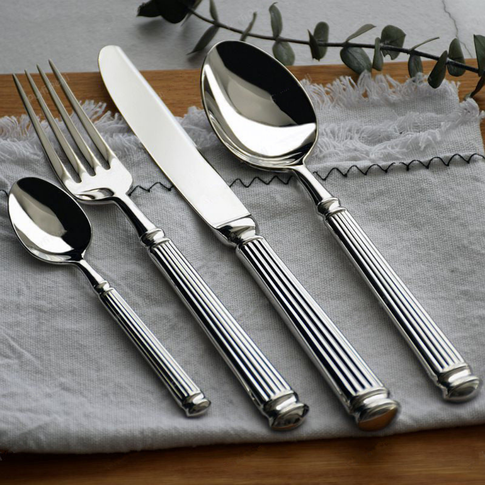Five pieces of mirror polished fluted cutlery laid on grey linen over a wooden board, with a eucalyptus sprig behind