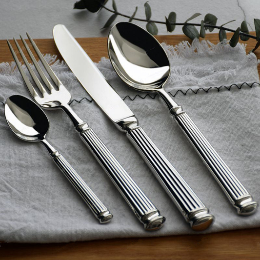 Five pieces of mirror polished fluted cutlery laid on grey linen over a wooden board, with a eucalyptus sprig behind