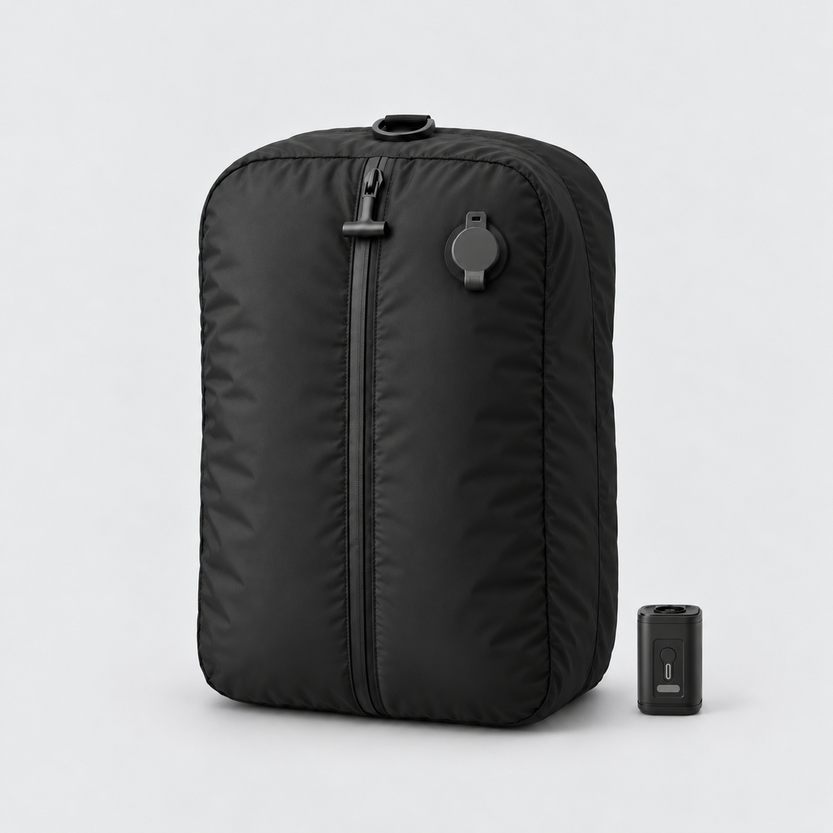 Black vacuum compression travel bag standing upright beside its square USB-C pump on a light grey background, with a flat ring handle on top and a dark grey capped air valve