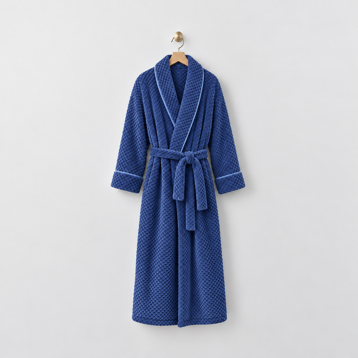 Long royal blue waffle-textured fleece bathrobe with a shawl collar and tied belt, hanging on a wooden hanger from a brass hook on a light gray wall
