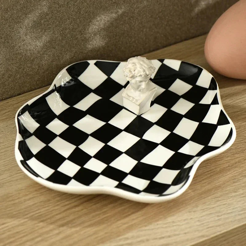 Black and white checkered ceramic trinket tray with a wavy scalloped edge, sitting on a wooden shelf in daylight