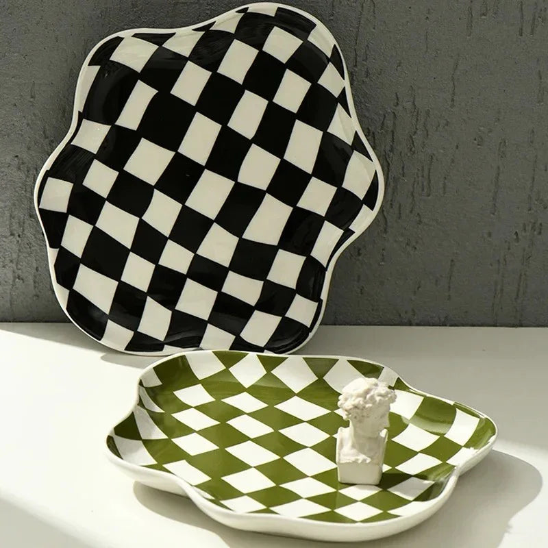 Black checkered ceramic tray propped against a grey wall behind a green checkered tray lying flat, both with the same wavy scalloped edge