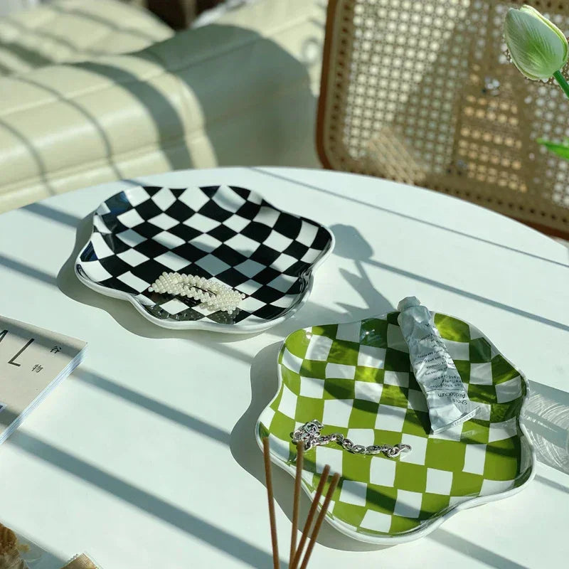 Two checkered ceramic trays in bright window light, a black tray holding a pearl hair clip and a green tray holding a chain bracelet