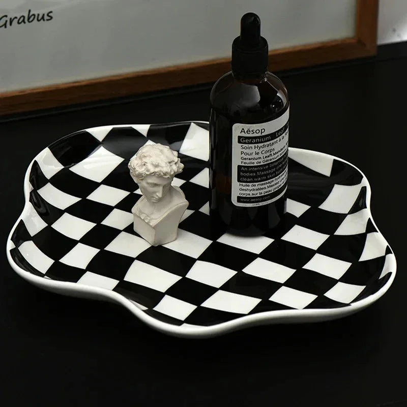 Black and white checkered ceramic tray on a dark surface, its glossy glaze catching the light, holding a small bottle and a plaster bust
