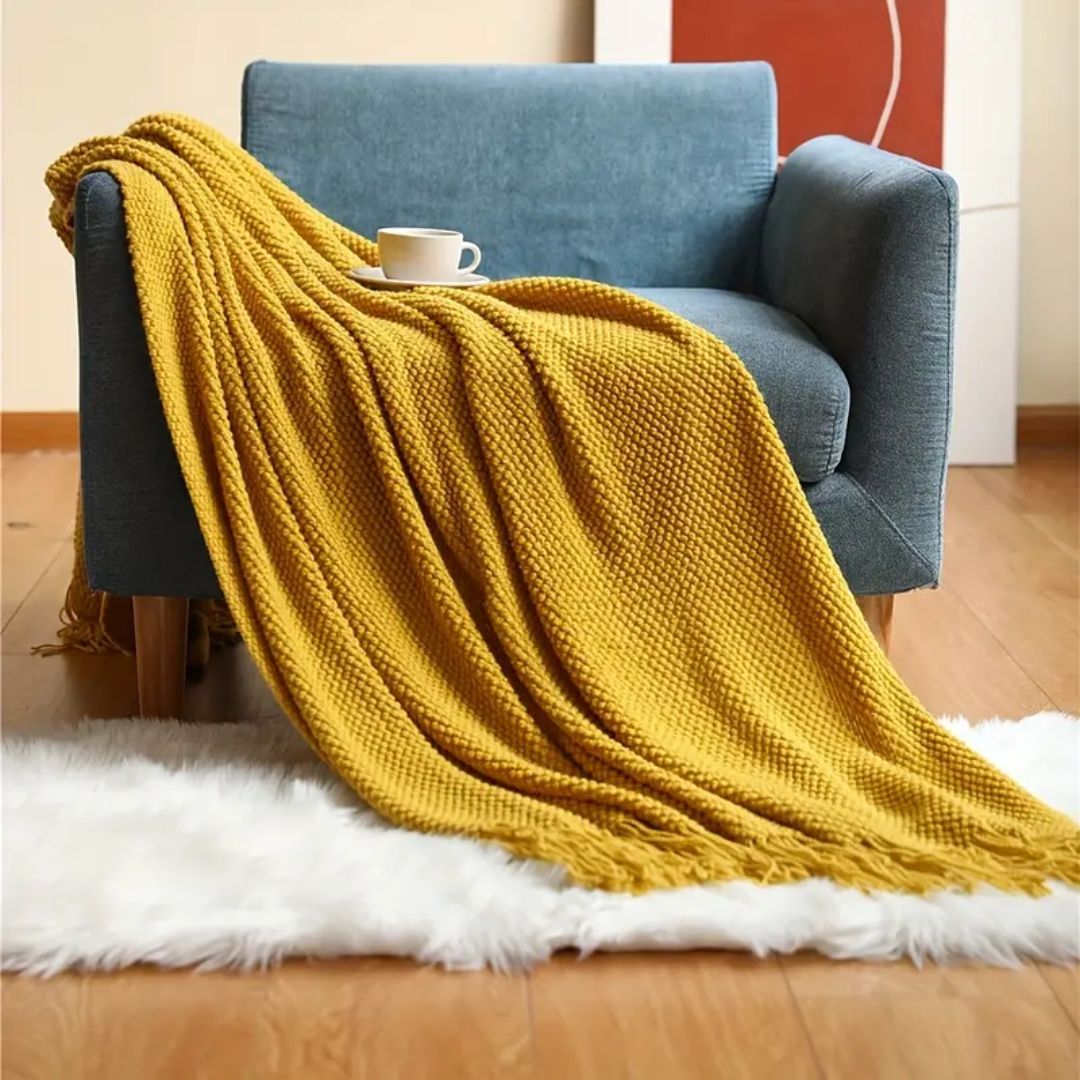 Mustard pebble knit throw blanket draped over a blue armchair with tassels