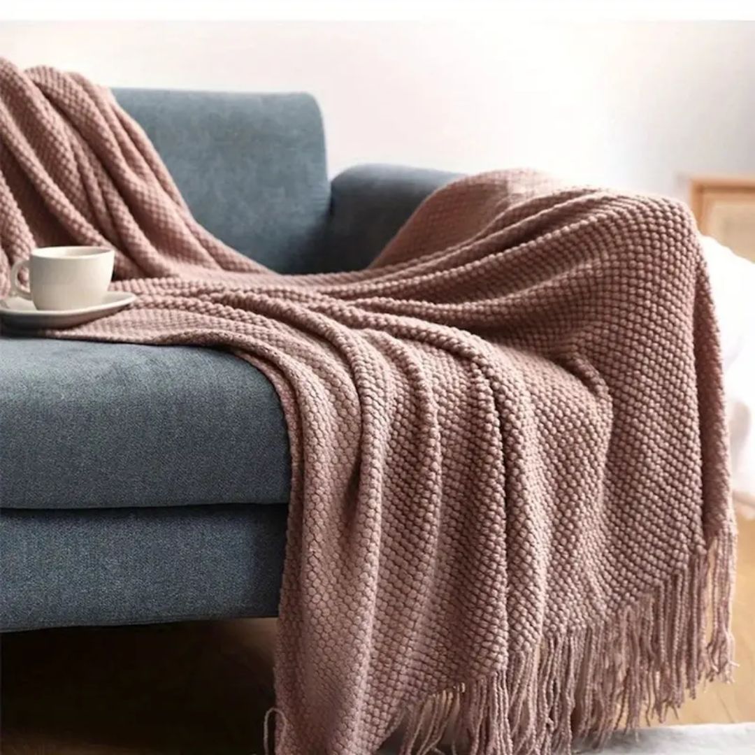 Dusty pink pebble knit throw blanket draped over a blue sofa with tassels