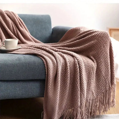 Dusty pink pebble knit throw blanket draped over a blue sofa with tassels