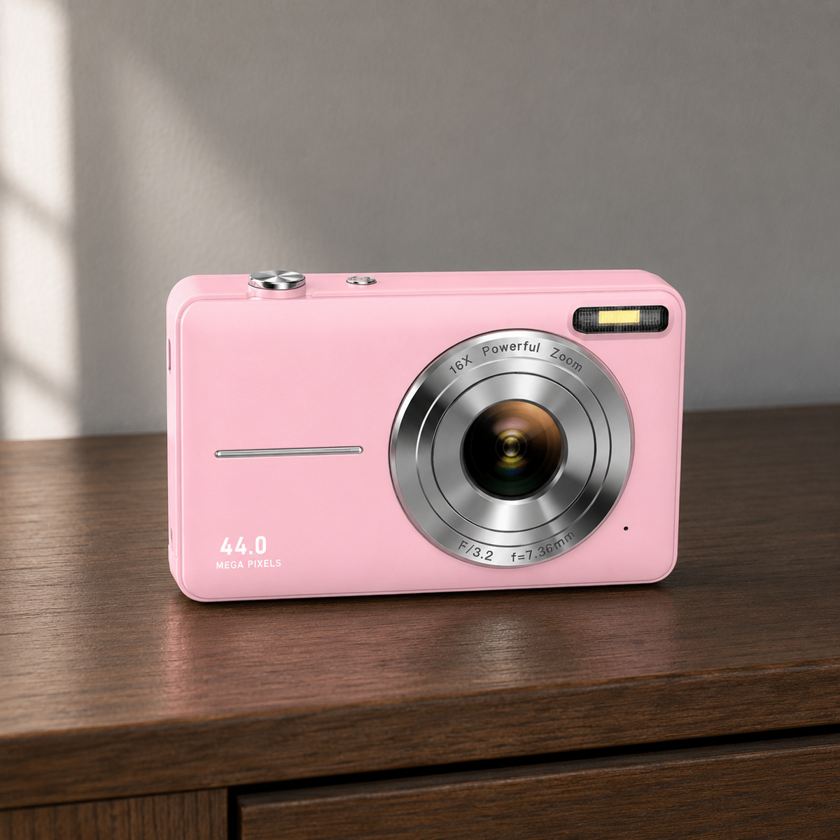Pink compact digital camera standing on a dark wood chest of drawers in a daylit bedroom, seen from the front at a slight angle, with the silver lens ring and 44.0 MEGA PIXELS on the lower left of the body
