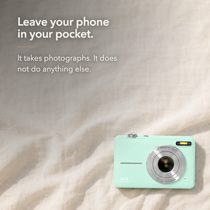 Text card. Green compact digital camera lying face up on a cream linen cloth photographed from directly above, headed Leave your phone in your pocket, above the line It takes photographs. It does not do anything else