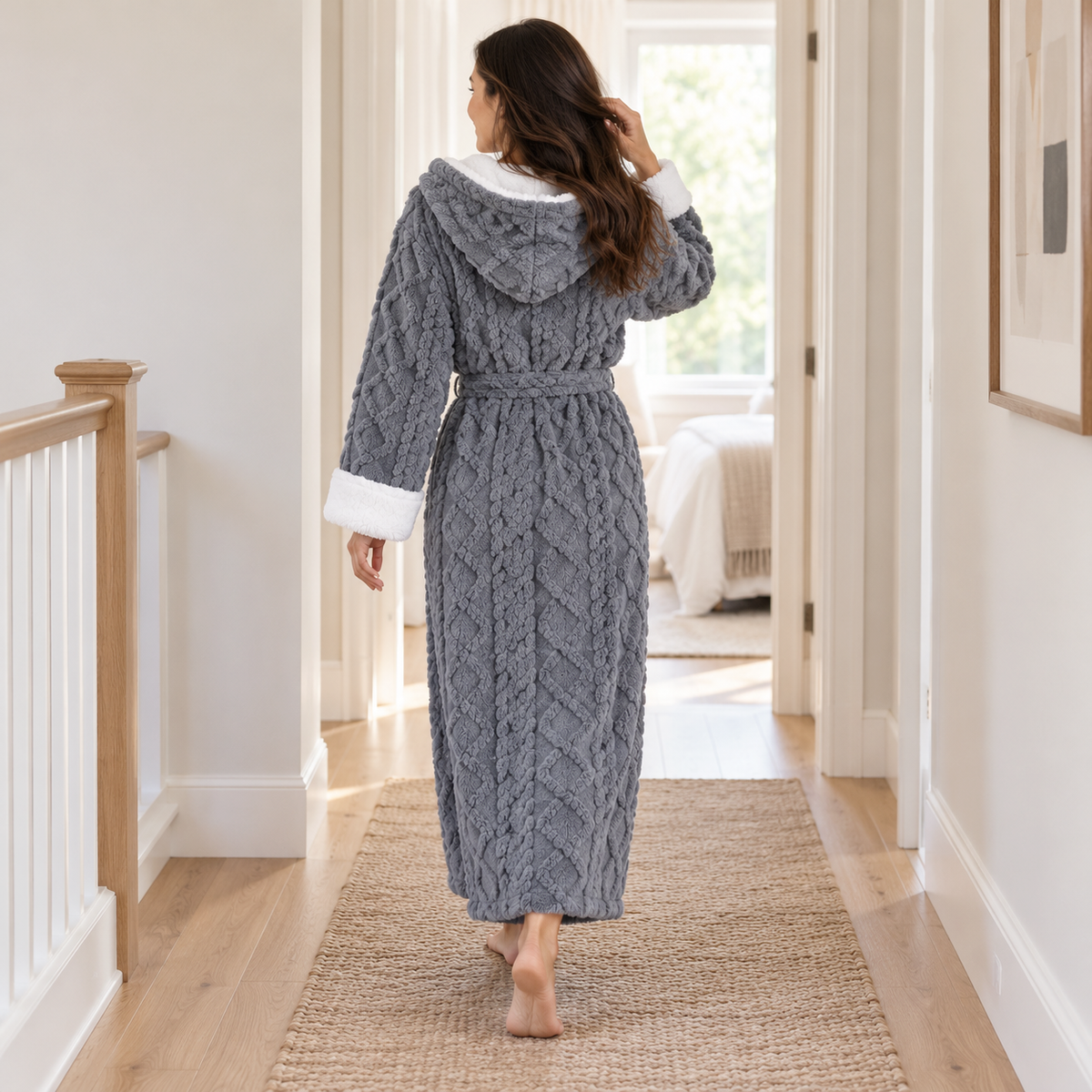 Woman in a gray plush bathrobe walking away down a hallway, showing the hood and the cable pattern on the back