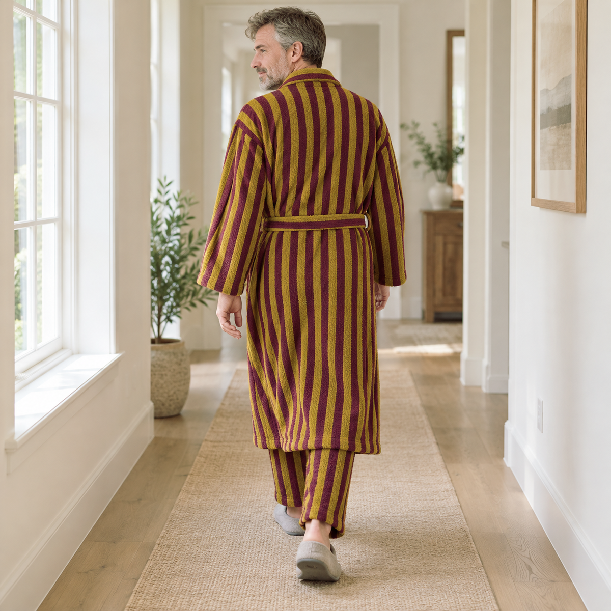 Back view of a man walking down a hallway in a long mustard and burgundy striped fleece bathrobe and matching pants