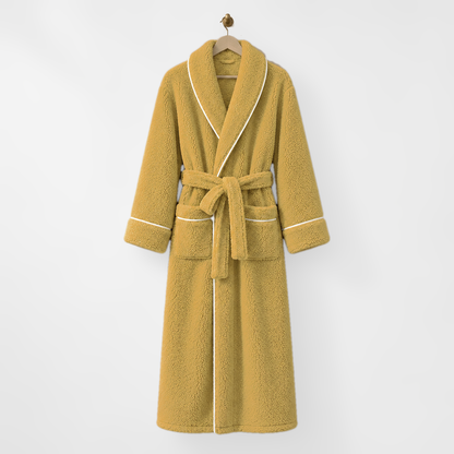 Long mustard fleece bathrobe with white piping, hanging on a wooden hanger from a brass hook on a light grey wall