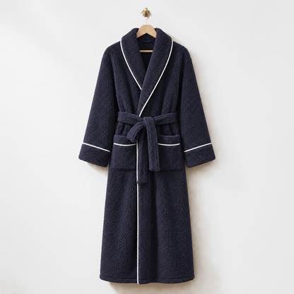 Long navy fleece bathrobe with white piping and a tied belt, hanging on a wooden hanger from a brass hook on a light grey wall