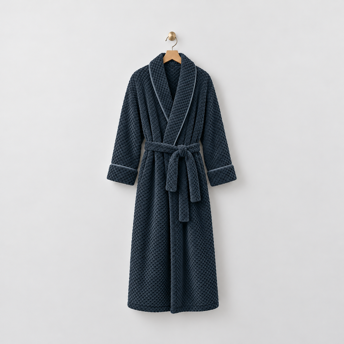 Long navy waffle-textured fleece bathrobe with a shawl collar and tied belt, hanging on a wooden hanger from a brass hook on a light gray wall