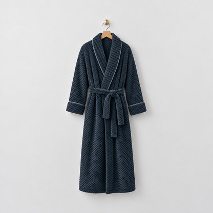 Long navy waffle-textured fleece bathrobe with a shawl collar and tied belt, hanging on a wooden hanger from a brass hook on a light gray wall