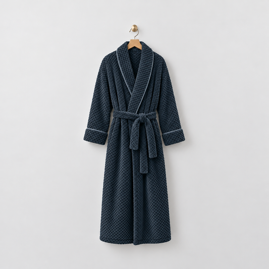 Long navy waffle-textured fleece bathrobe with a shawl collar and tied belt, hanging on a wooden hanger from a brass hook on a light gray wall