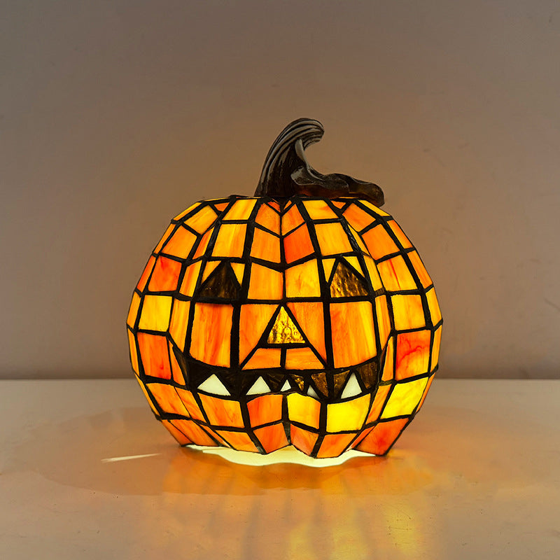 Mosaic jack-o'-lantern table lamp lit on a table, orange and amber mosaic glowing around a grinning face