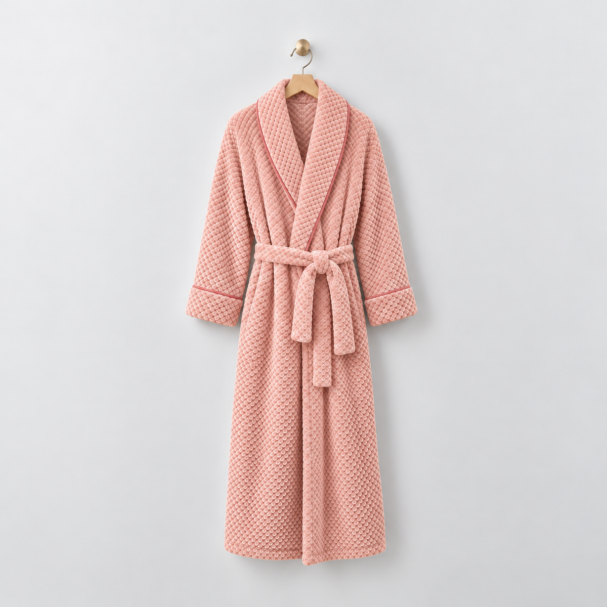 Long pink waffle-textured fleece bathrobe with a shawl collar and tied belt, hanging on a wooden hanger from a brass hook on a light gray wall