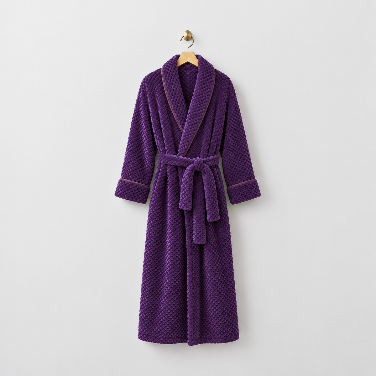 Long purple waffle-textured fleece bathrobe with a shawl collar and tied belt, hanging on a wooden hanger from a brass hook on a light gray wall