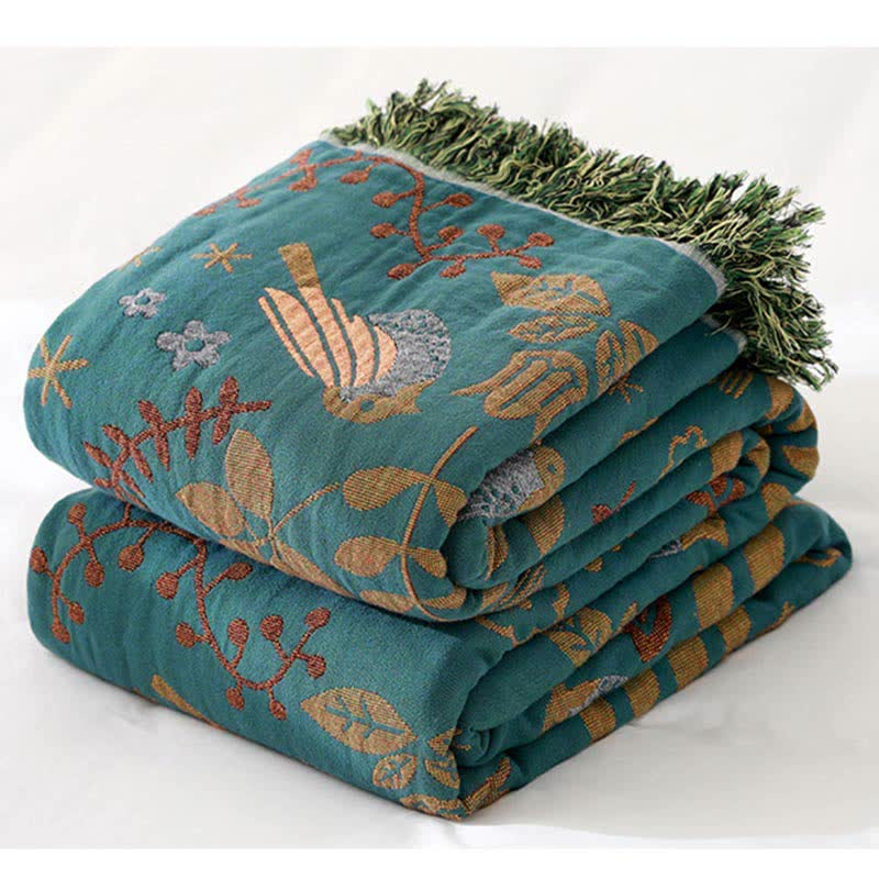 The teal bird throw folded in a stack, showing a golden bird with a speckled gray body, rust red sprigs and the green fringe
