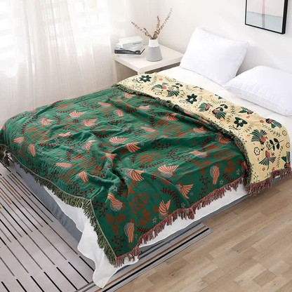 The green version of the bird throw spread across a made bed, with salmon pink birds and burgundy sprigs, and its cream side with dark green birds folded back at the pillows