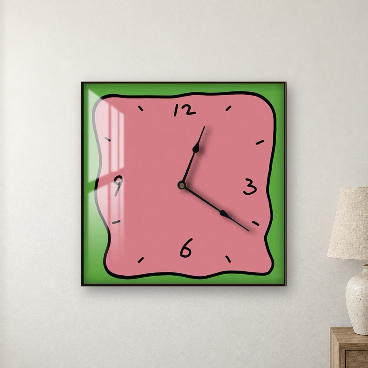 Square wall clock with a wavy doodle-style outline, a pink face and a green border, hanging centred on a warm white wall beside a bedside lamp