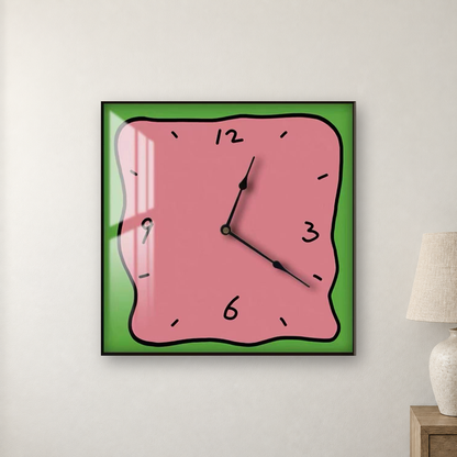 Square wall clock with a wavy doodle-style outline, a pink face and a green border, hanging centred on a warm white wall beside a bedside lamp