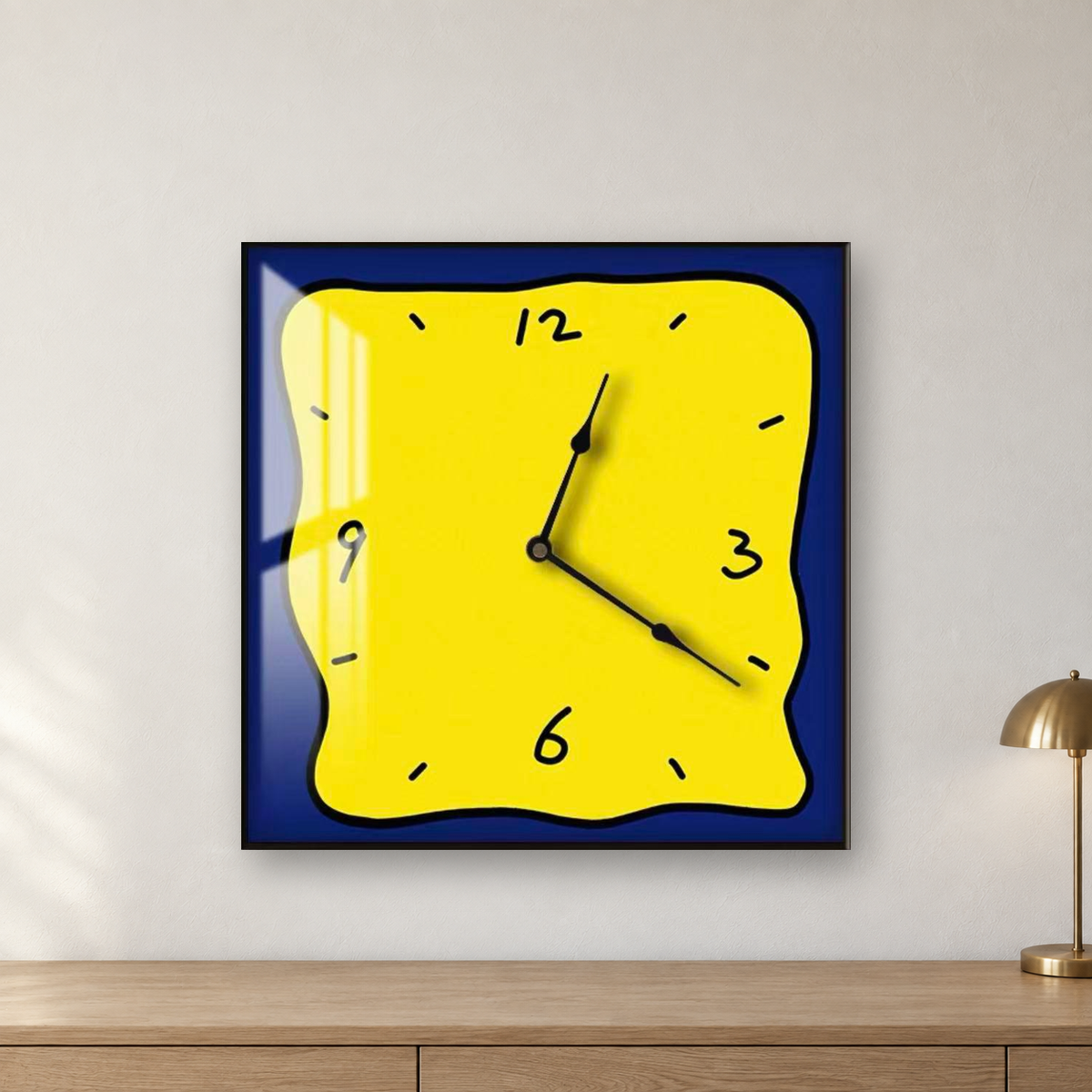 Square wall clock with a wavy doodle-style outline, a yellow face and a navy border, hanging centred on a white plaster wall above an oak cabinet with a brass lamp