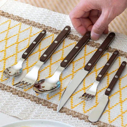 Brown wood-grain look handled six piece flatware set on a woven yellow table runner, a hand touching one piece