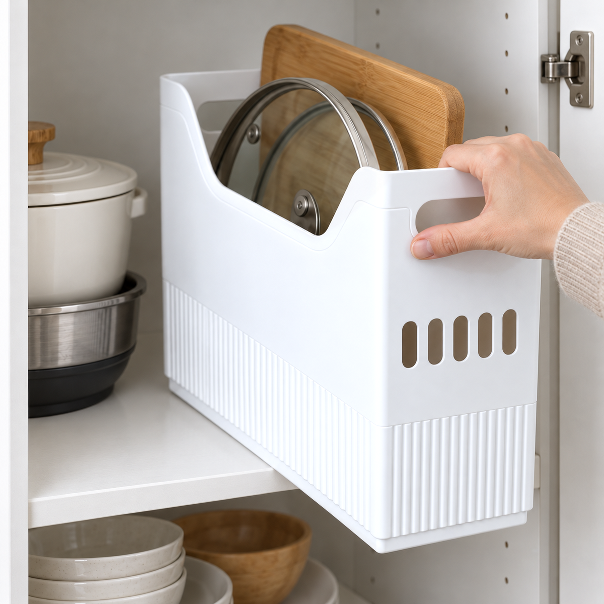 A hand gripping the top edge of the end of a white storage bin of pan lids and a cutting board, drawing it forward off a kitchen cabinet shelf