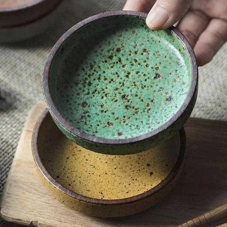 Green speckled ceramic dipping bowl held above a yellow speckled dish on a wooden board, both with a brown rim