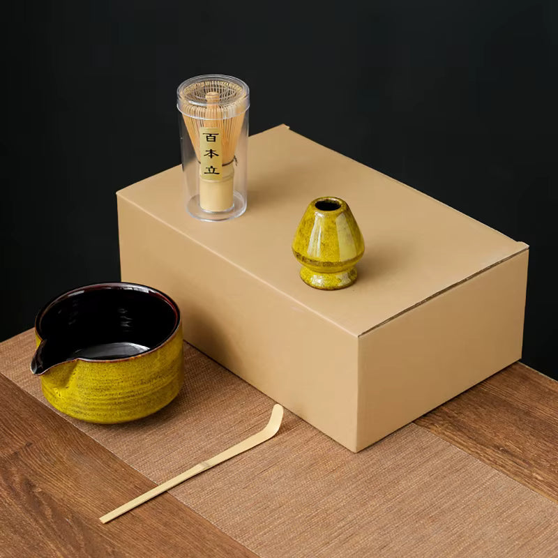 Yellow four-piece matcha kit: spouted ceramic bowl, bamboo scoop, whisk holder and bamboo whisk, shown on a plain carton