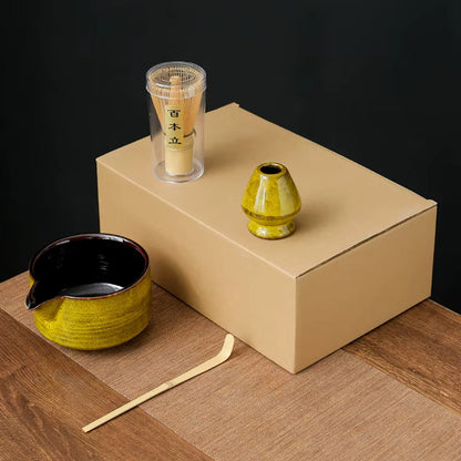 Yellow four-piece matcha kit: spouted ceramic bowl, bamboo scoop, whisk holder and bamboo whisk, shown on a plain carton