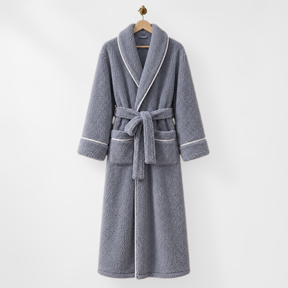 Long slate blue-grey fleece bathrobe with white piping, hanging on a wooden hanger from a brass hook on a light grey wall