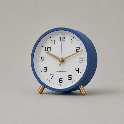 Analog alarm clock in deep navy blue, studio shot on warm grey, showing the white dial, gold hands and gold-toned feet