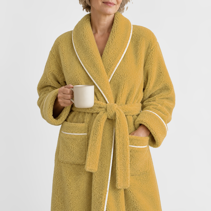 Close view of a woman in a mustard fleece bathrobe with white piping, holding a cream mug, on a light grey backdrop