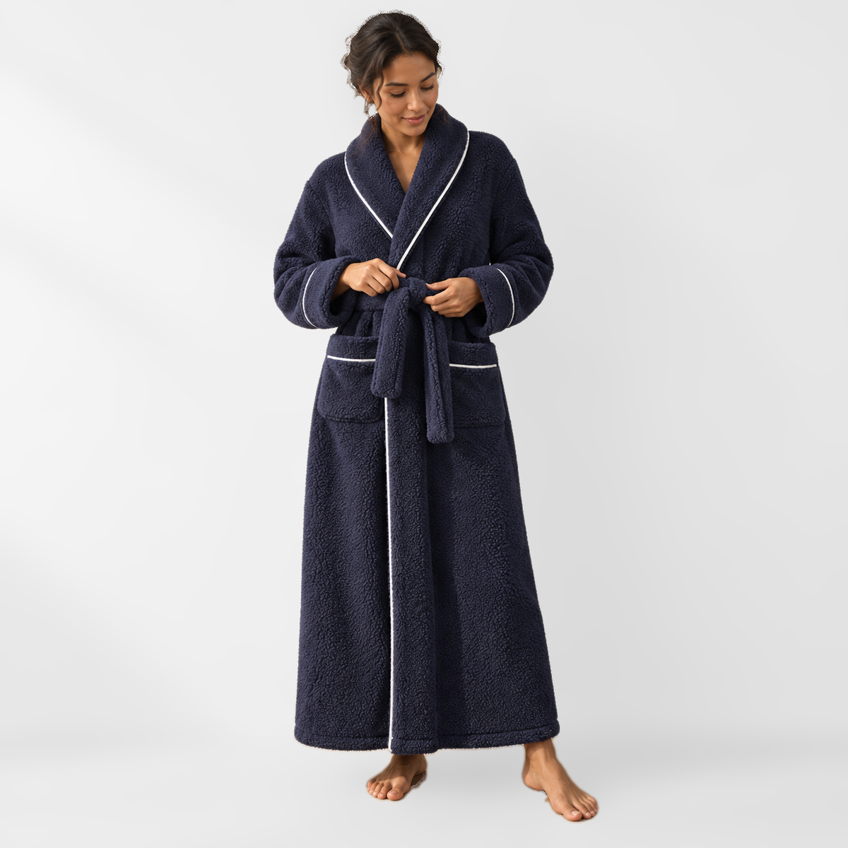 Woman tying the belt of an ankle-length navy fleece bathrobe with white piping, on a light grey backdrop